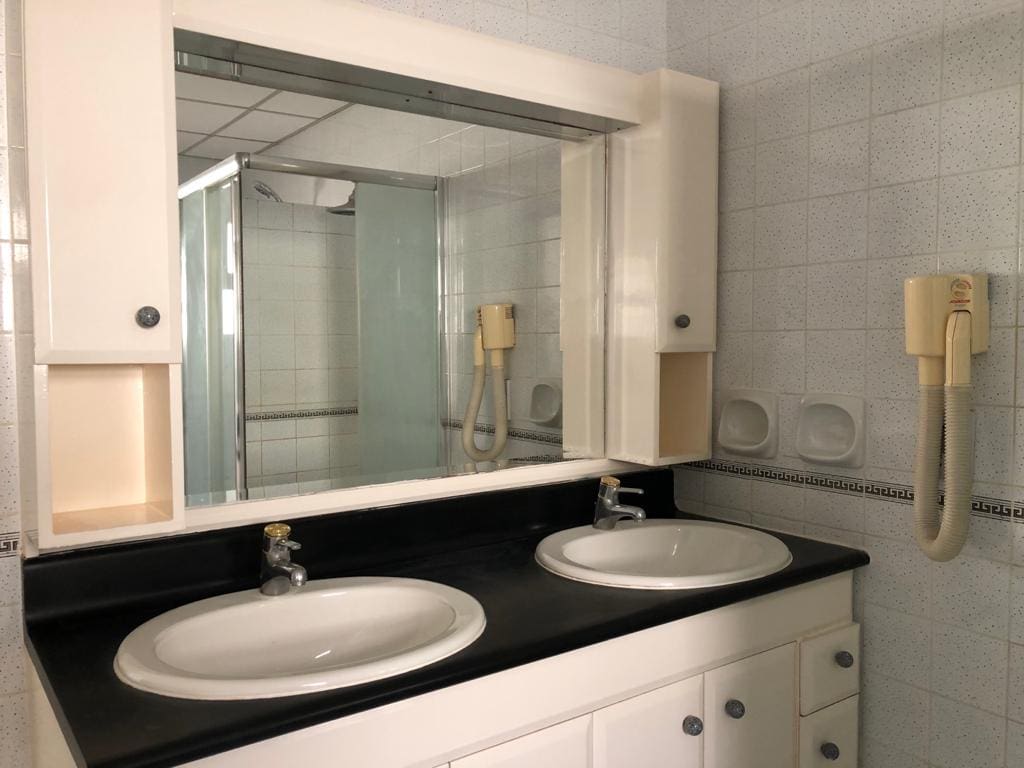 A bathroom with a double sink vanity, wall-mounted hairdryer, and a large mirror. The room features white tiles and a corner shower with glass doors.