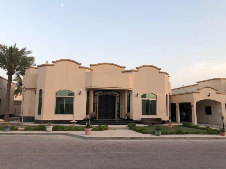 A two-story beige house with curved architectural elements, a large black front door, and a garage on the right. The house has symmetrical greenery and potted plants along the front entrance.