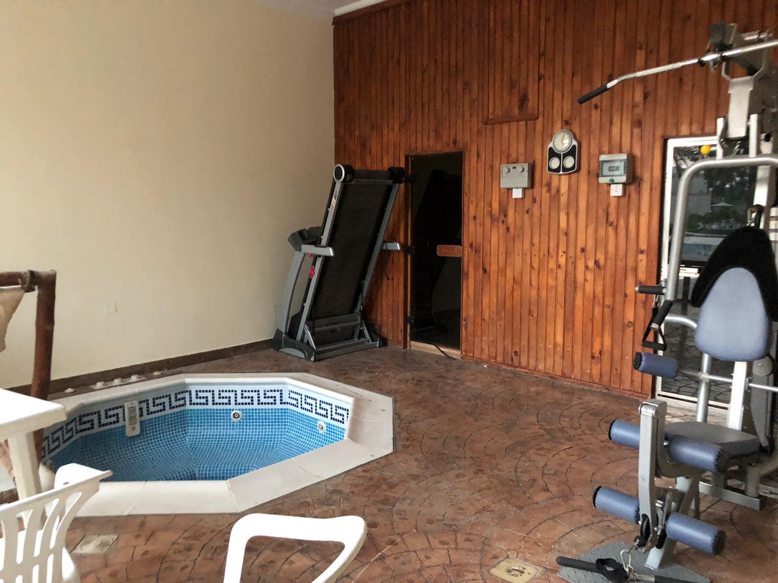 A small indoor gym with exercise equipment, a treadmill, and a corner hot tub surrounded by a tiled floor and wooden wall panels. A white plastic chair is in the foreground.