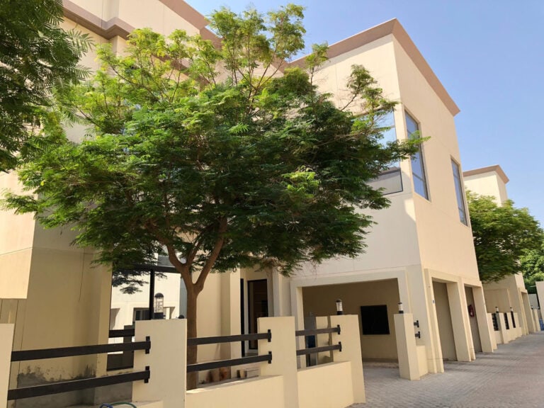 A modern, beige-colored townhouse with large windows, a small front yard, and a tree providing shade. The building has a minimalist design with a clean exterior and a paved walkway.