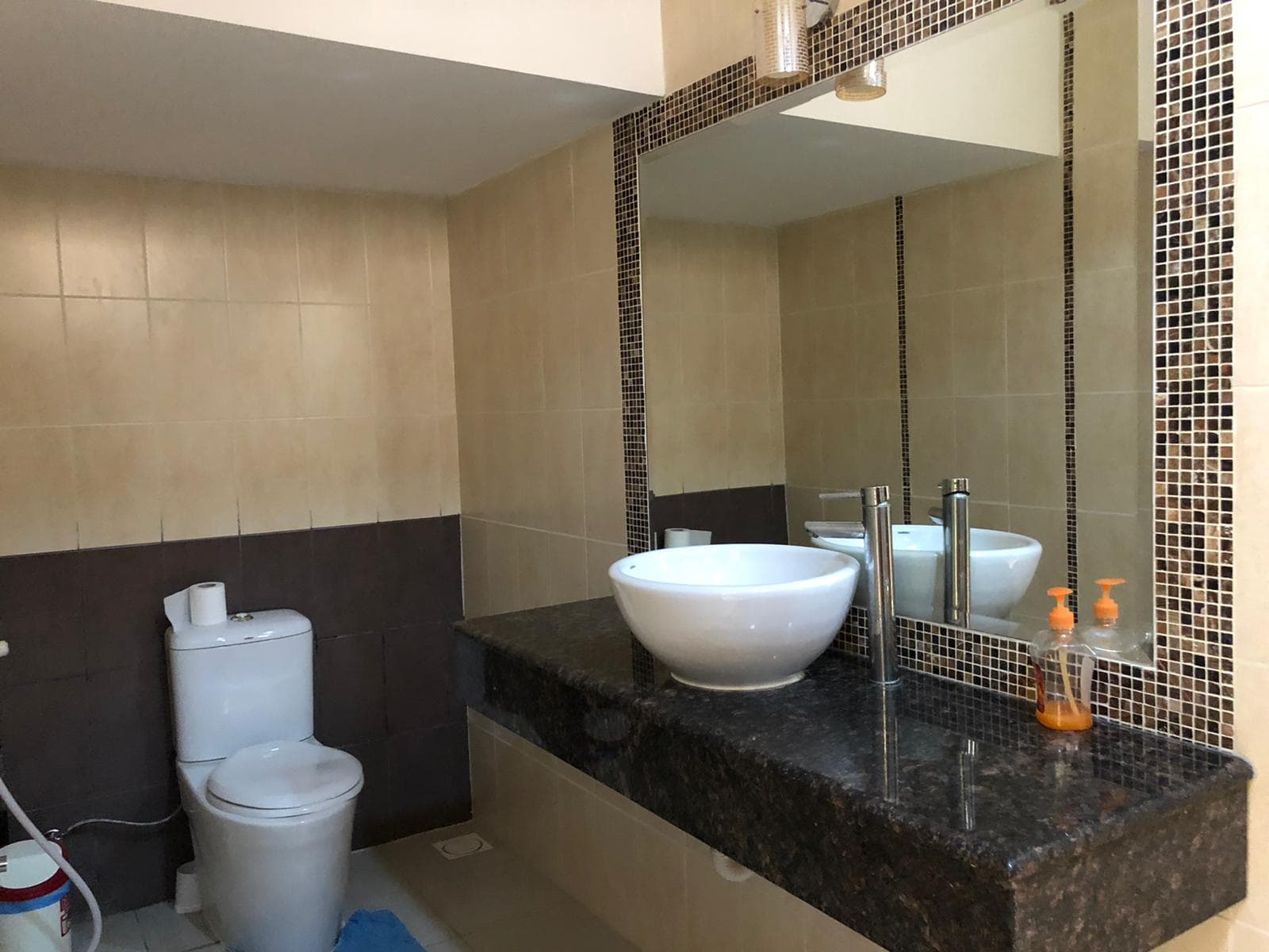 A bathroom with beige tiles, a large mirror, a white toilet, a vessel sink on a granite countertop, and a soap dispenser.