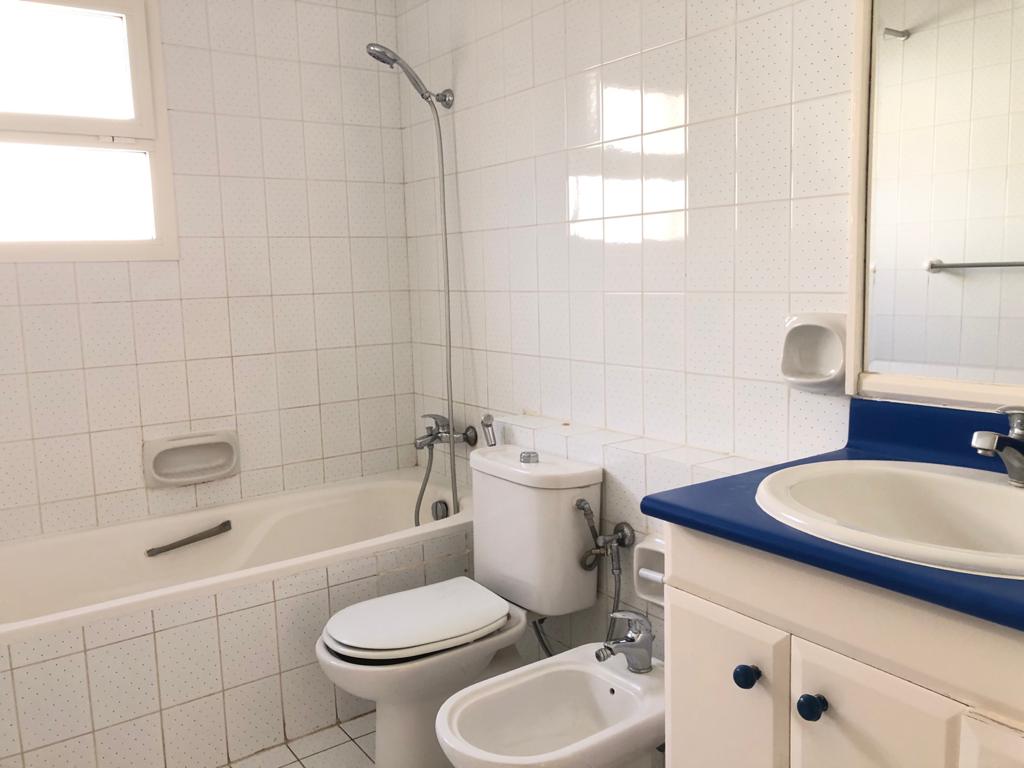 A clean, white-tiled bathroom with a bathtub-shower combo, toilet, bidet, and a sink with a blue countertop and a mirror above it.