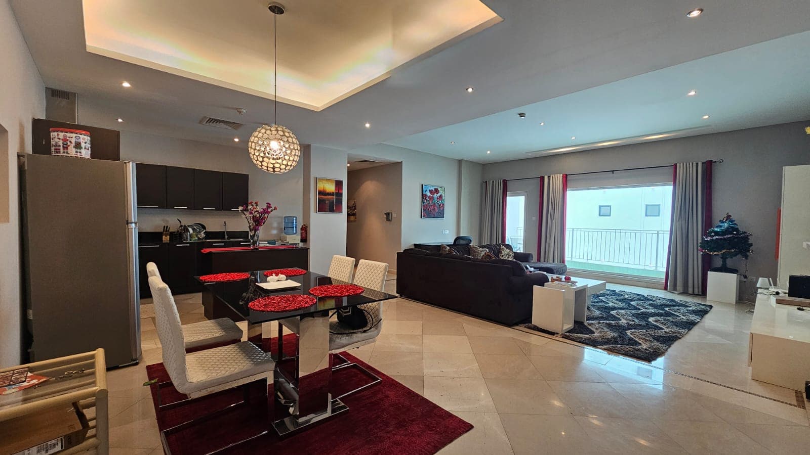 Modern living room and dining area with an open floor plan. The space includes a dining table, kitchen, couches, TV, large window with balcony, and various decorations.