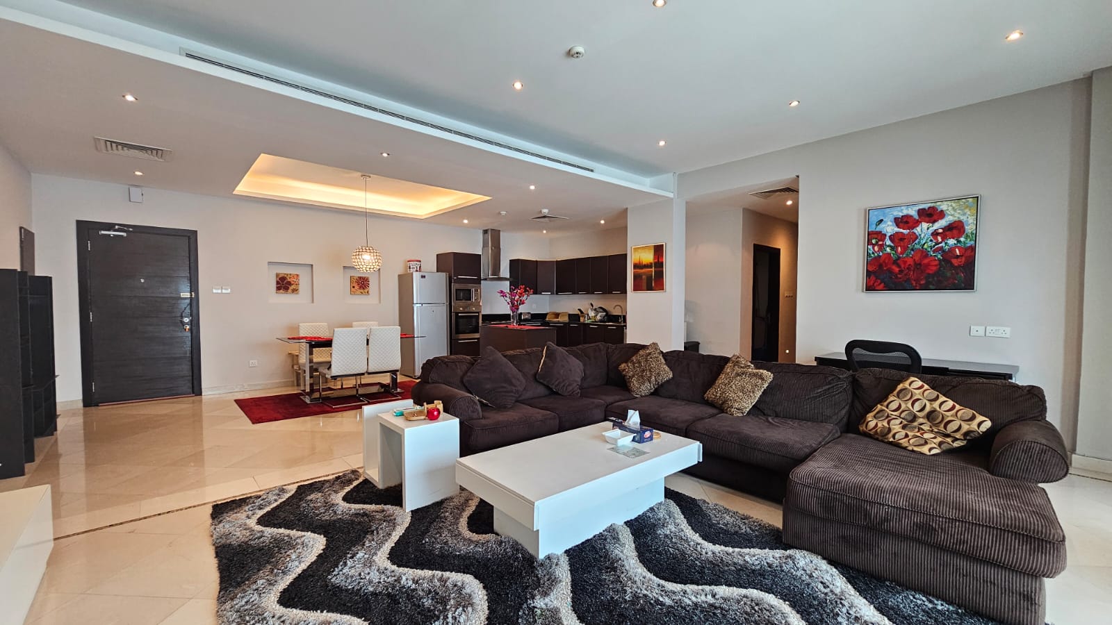 A modern living room with a dark sectional sofa, a white coffee table, and a patterned rug. The room extends into an open-plan kitchen and dining area, featuring a table with white chairs.