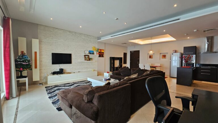 Spacious living room with a brown sectional sofa, wall-mounted TV, Christmas tree, and dining area. Modern kitchen in the background with stainless steel appliances and white cabinetry.