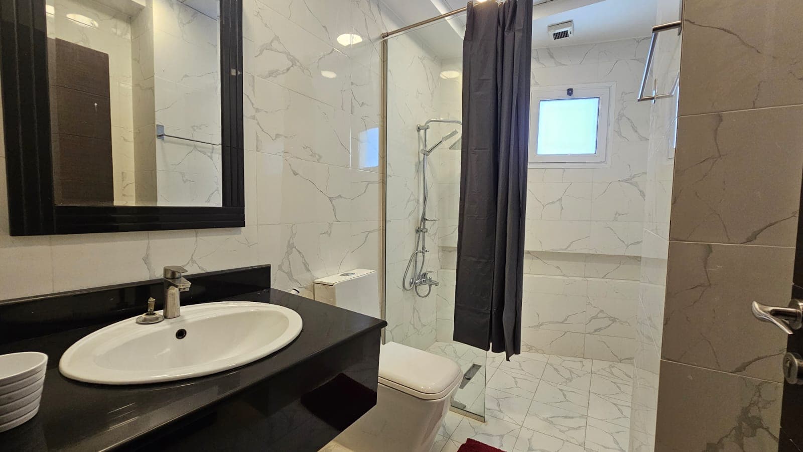 A bathroom featuring a sink with a mirror, a toilet, and a glass-enclosed shower area with a curtain. The walls and floor are tiled with white and gray patterns, and a small window is near the shower.