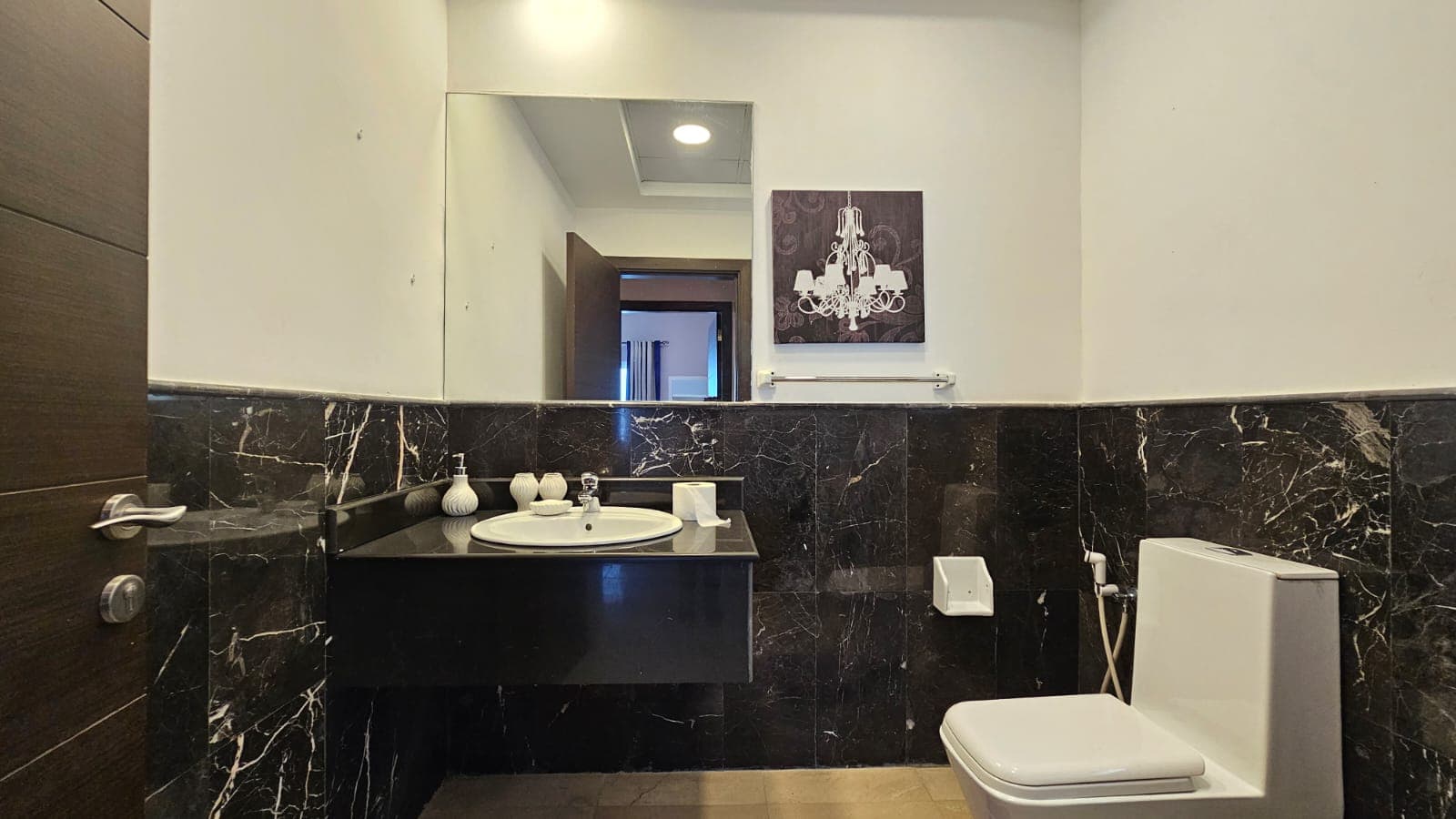 A modern bathroom features a wall-mounted sink, a toilet, a mirror, black marble wall tiles, and a chandelier artwork on the wall. A door is partially open, revealing another room in the background.