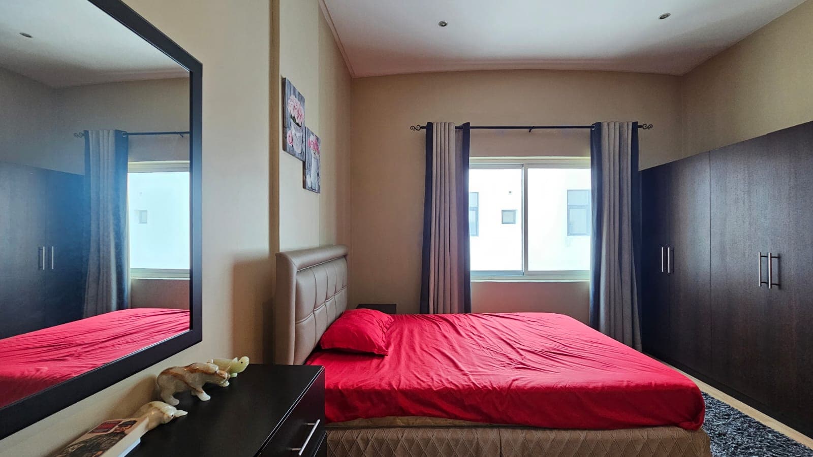 A bedroom with a red bedspread, dark wooden wardrobes, a window with curtains, and a mirror reflecting the room. There is a bedside table with items on it and framed pictures on the wall.