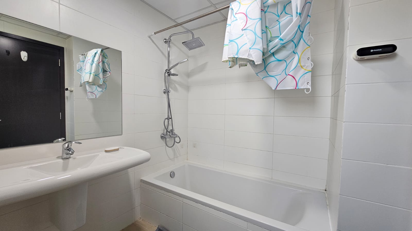 A bathroom with white tiled walls, a bathtub with a showerhead, a white sink with a mirror above it, and a colorful shower curtain. A small electronic device is mounted on the right wall.