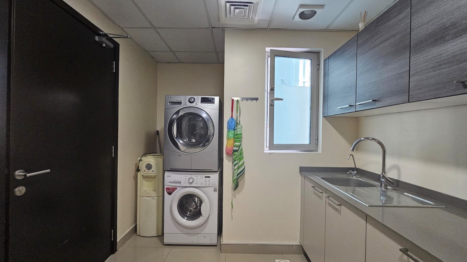 A laundry room with a stackable washer and dryer, a sink with a faucet, overhead cabinets, and a window. Cleaning supplies are also present.