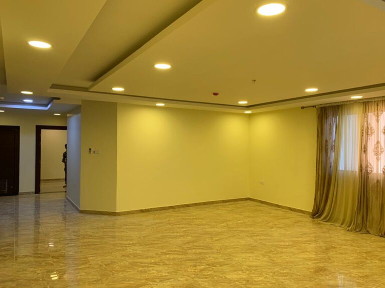 A spacious, empty room with marble tile flooring, recessed ceiling lights, and a large window with sheer curtains, creating a bright and open atmosphere.