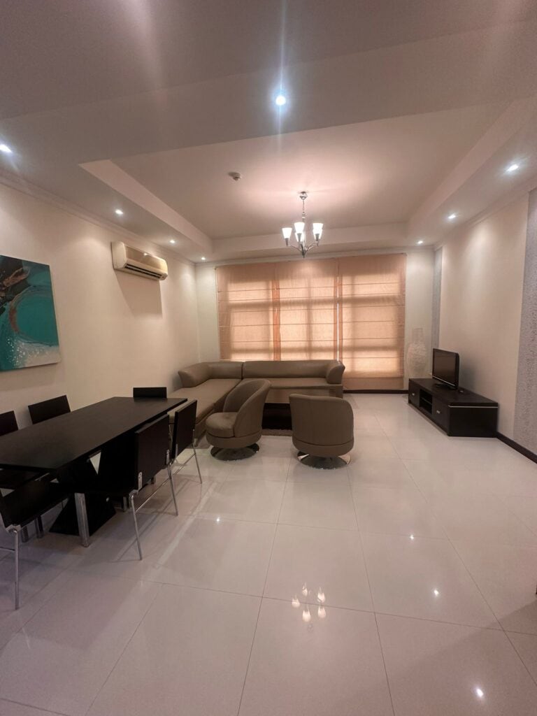 A modern living room with a dining table, chairs, a sectional sofa, armchairs, a TV, and a chandelier. The room is well-lit with natural light from the large window and has white tile flooring.