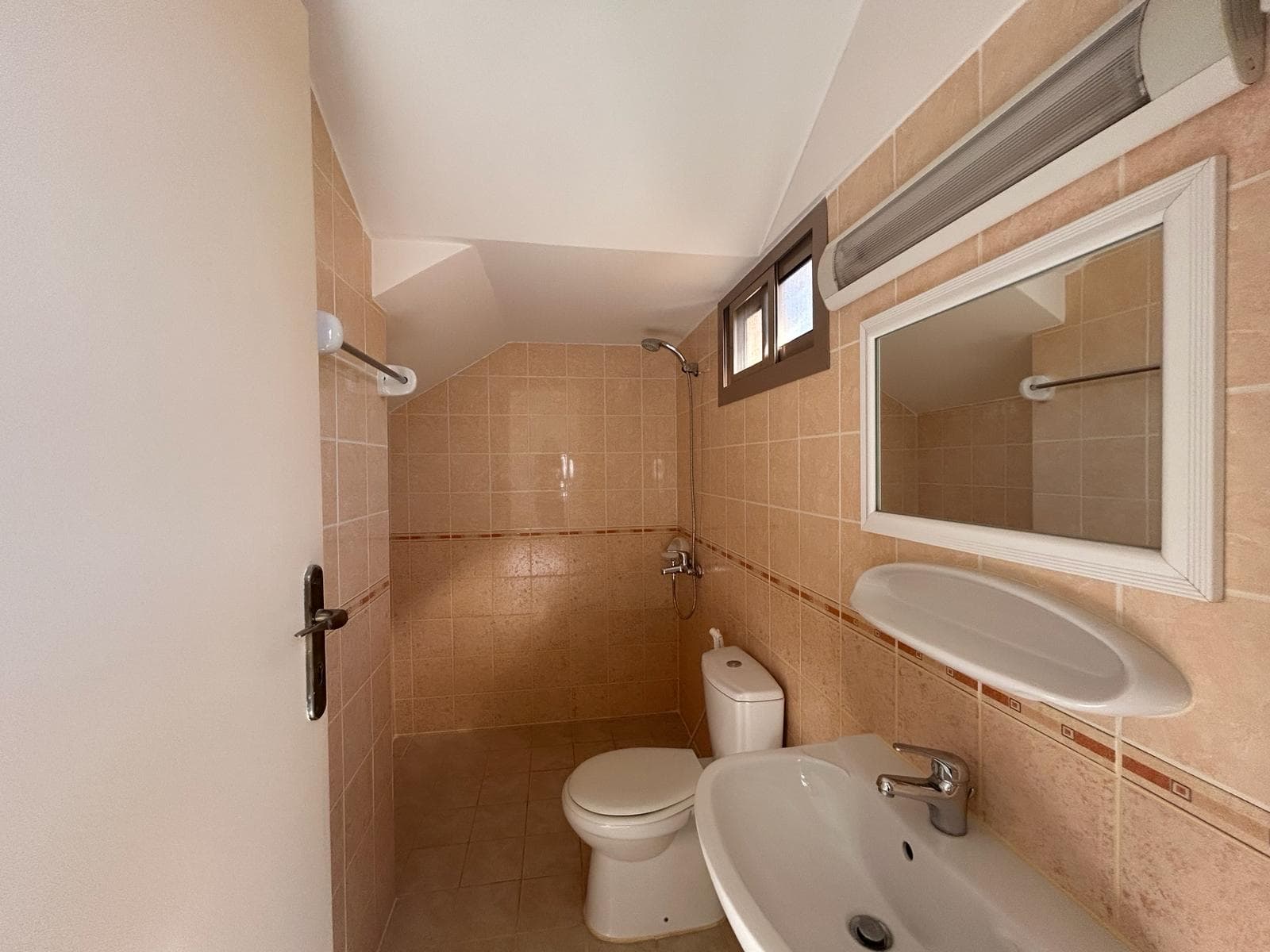 Small bathroom with beige tiled walls, featuring a toilet, sink, mirror, and overhead light. A narrow window is placed above the toilet, and the door is partially open.