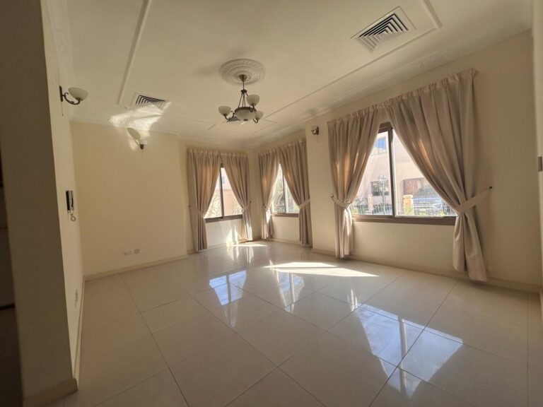 A sunlit room with large windows covered by beige curtains, cream-colored tiled flooring, and a ceiling with multiple light fixtures.