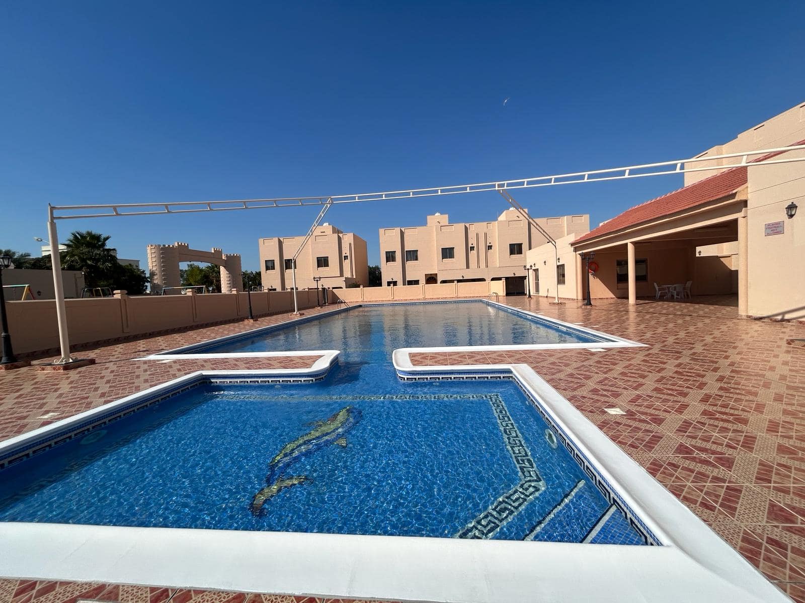 A residential swimming pool area with a main pool and a smaller shallow section. The tiled pool deck has scattered furniture, and rows of houses are seen in the background under a clear blue sky.