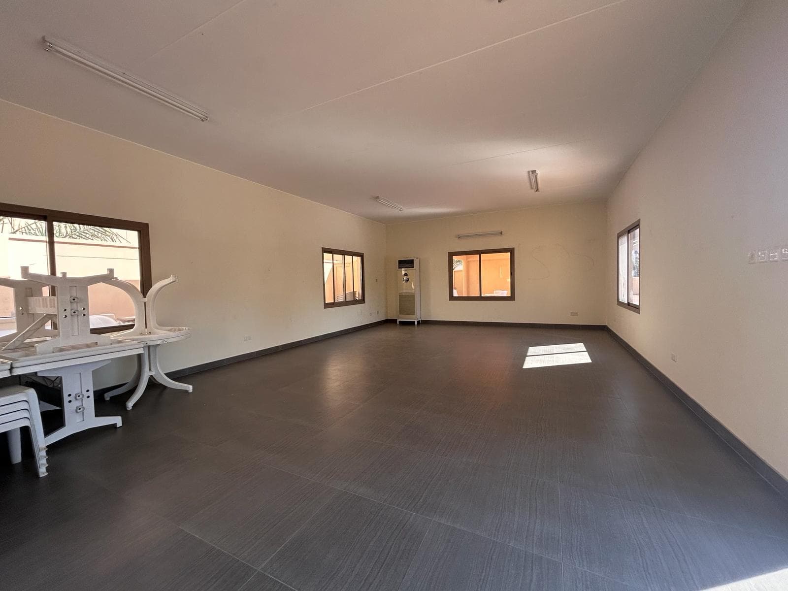 A spacious, empty room with tiled flooring, multiple windows, fluorescent lights, and stacked tables and chairs in one corner.