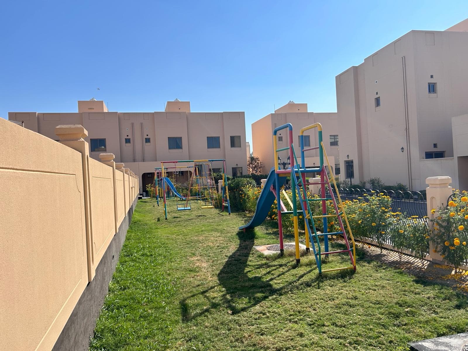 A small grassy playground with colorful climbing frames, slides, and swings is situated between beige residential buildings under a clear blue sky.