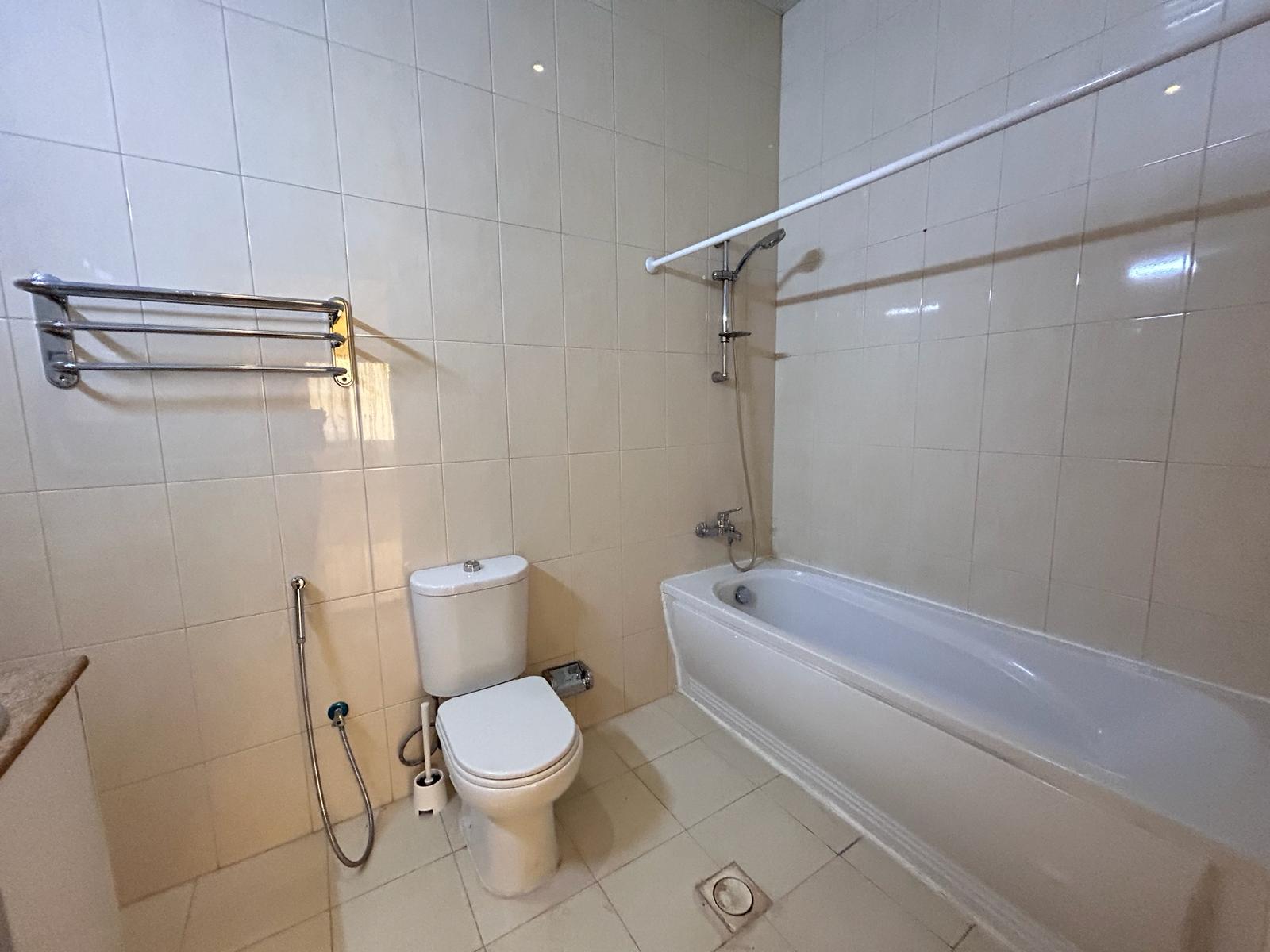 A bathroom with white tiles, featuring a toilet, a bidet sprayer, a bathtub with a shower overhead, and a towel rack.