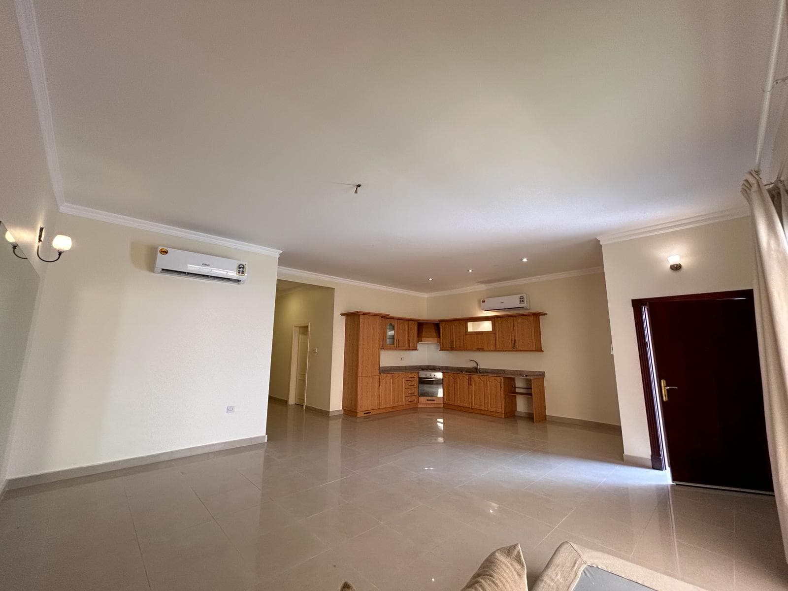 A spacious, unfurnished room with a tiled floor, featuring an open kitchen with wooden cabinets, two air conditioning units, a closed door, and light fixtures on the walls.