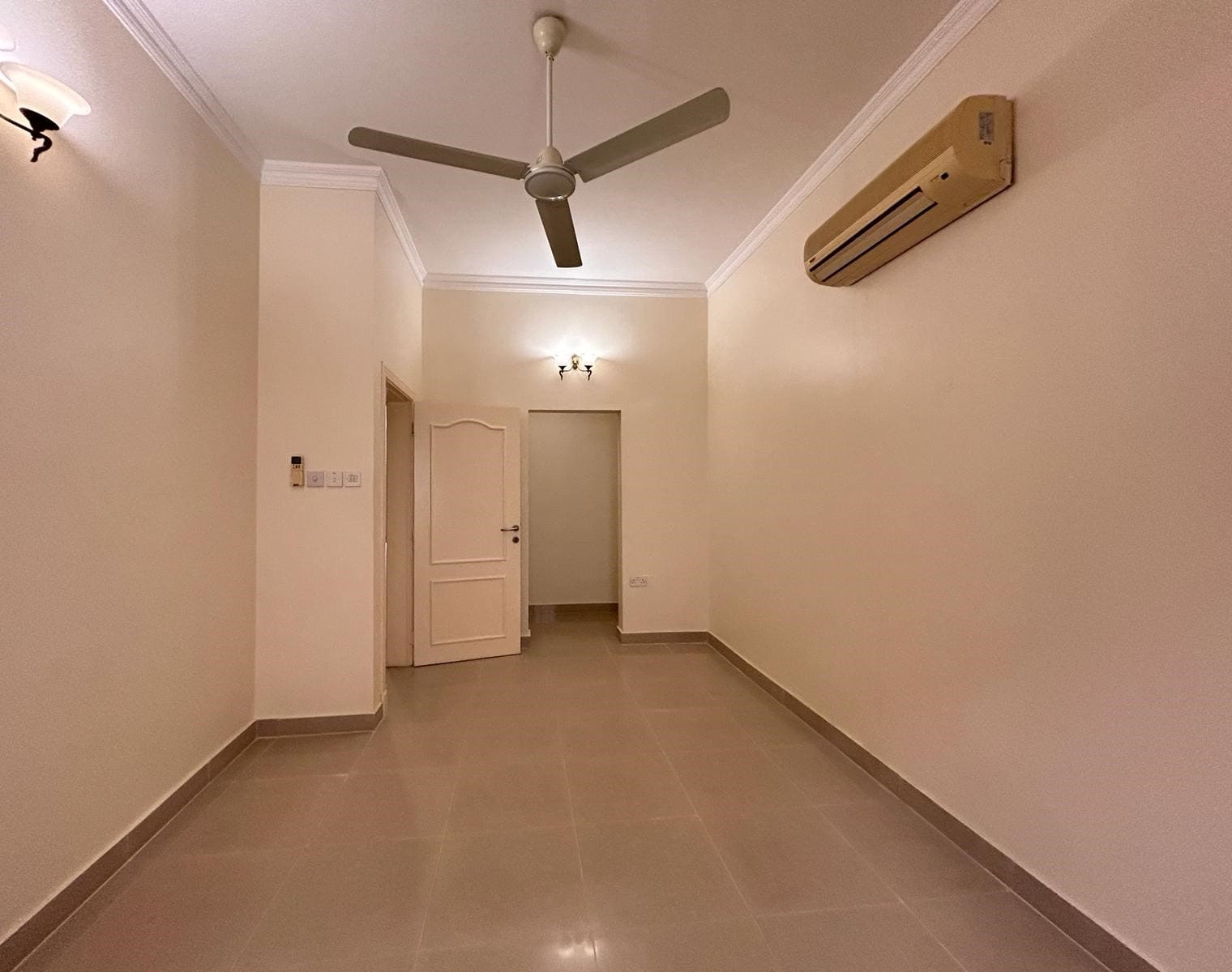 Empty room with tiled floor, beige walls, ceiling fan, air conditioner, and two wall-mounted light fixtures. Door visible in the back leads to another area.