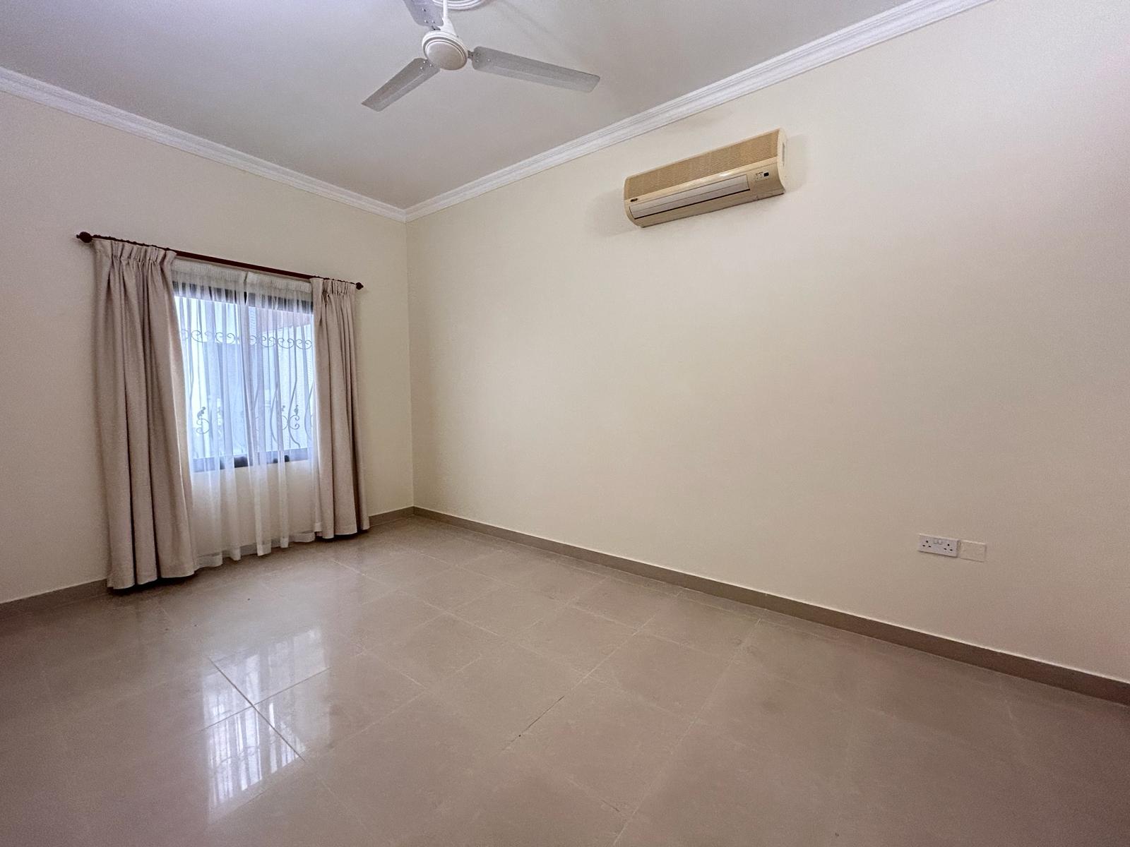 A spacious, empty room with beige walls, a ceiling fan, an air conditioner, a window with curtains, and a tiled floor.