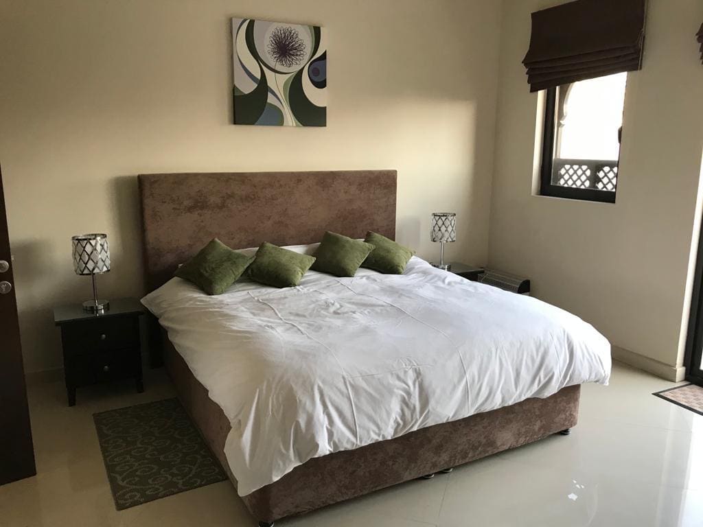 A neatly arranged bedroom featuring a large bed with a brown headboard, white bedding, green cushions, two bedside tables with lamps, a wall painting, and a window with brown blinds.