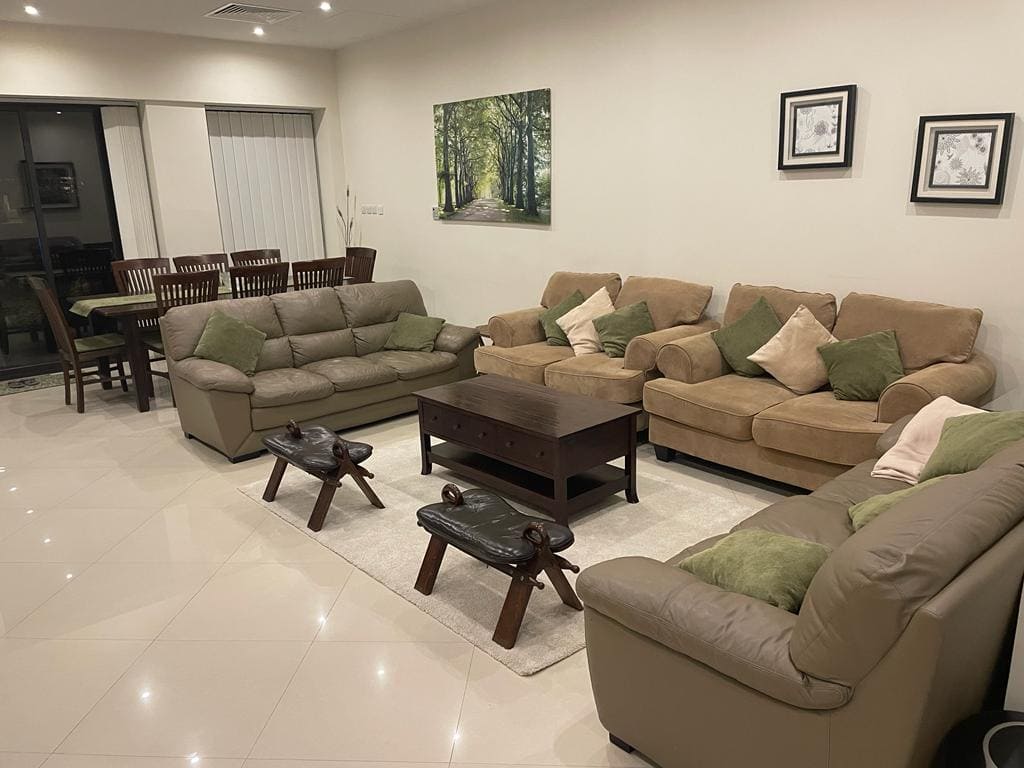 A spacious living room with two beige sofas, a brown coffee table, and a dining area in the background. The room is decorated with framed wall art and green cushions.