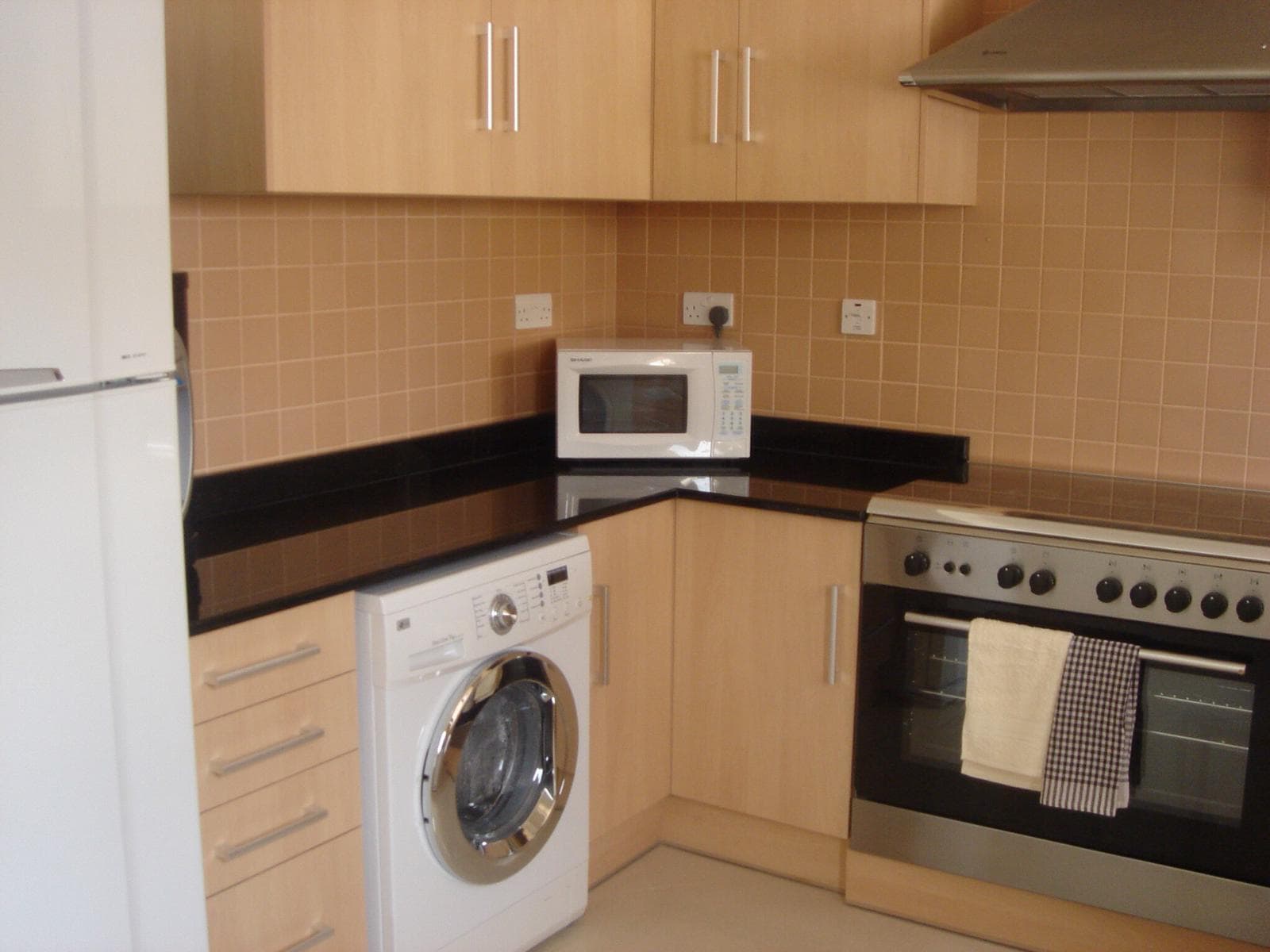 A modern kitchen featuring wooden cabinets, a black countertop, a washing machine, a microwave, a stove with an oven, and a towel hanging on the oven handle.