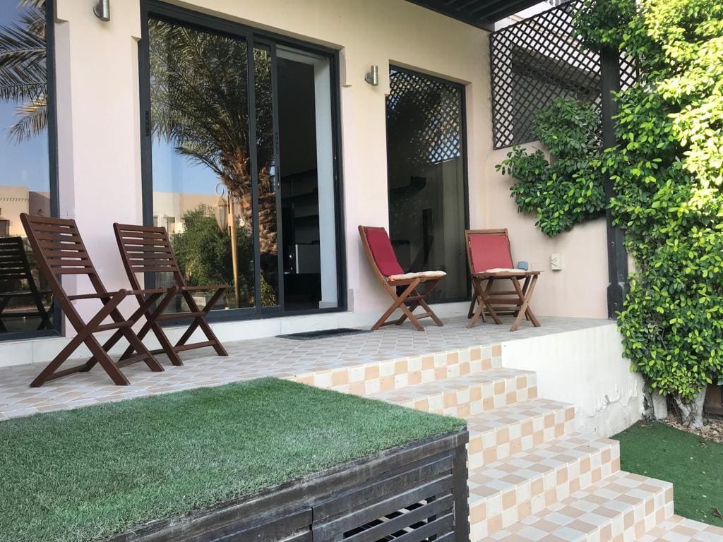 Outdoor patio with wooden chairs and a table on a tiled platform. The patio is adjacent to a building with glass doors and greenery on one side. Steps lead down to a small grassy area.