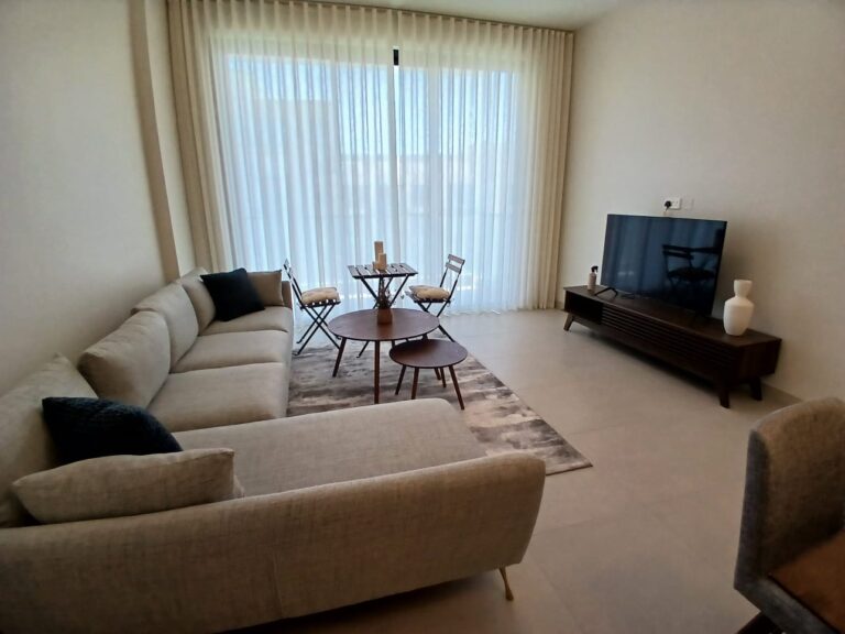 A living room with a beige sectional sofa, a flat-screen TV on a wooden stand, a small dining table with two chairs, a round coffee table, and floor-to-ceiling curtains covering a large window.