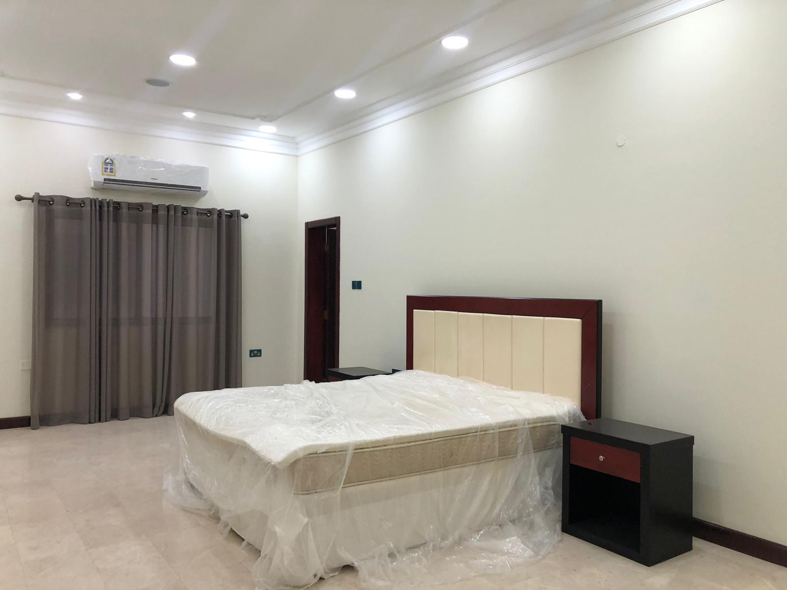 A bedroom with a double bed wrapped in plastic, two bedside tables, a window with closed grey curtains, and a wall-mounted air conditioner. The room has white walls and ceiling with recessed lighting.