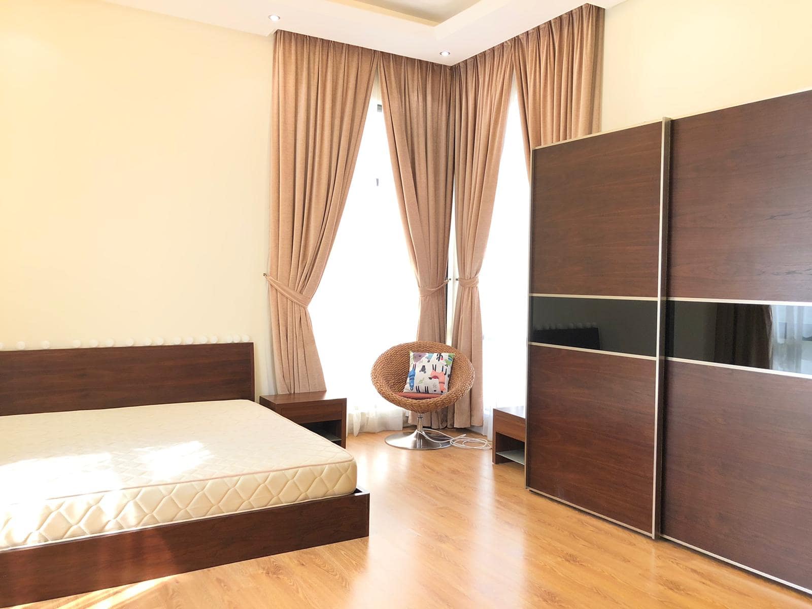 A bedroom with a wooden bed frame and matching nightstand, large windows with beige curtains, a cushioned chair, and a dark wood wardrobe. The room has light-colored walls and wooden flooring.