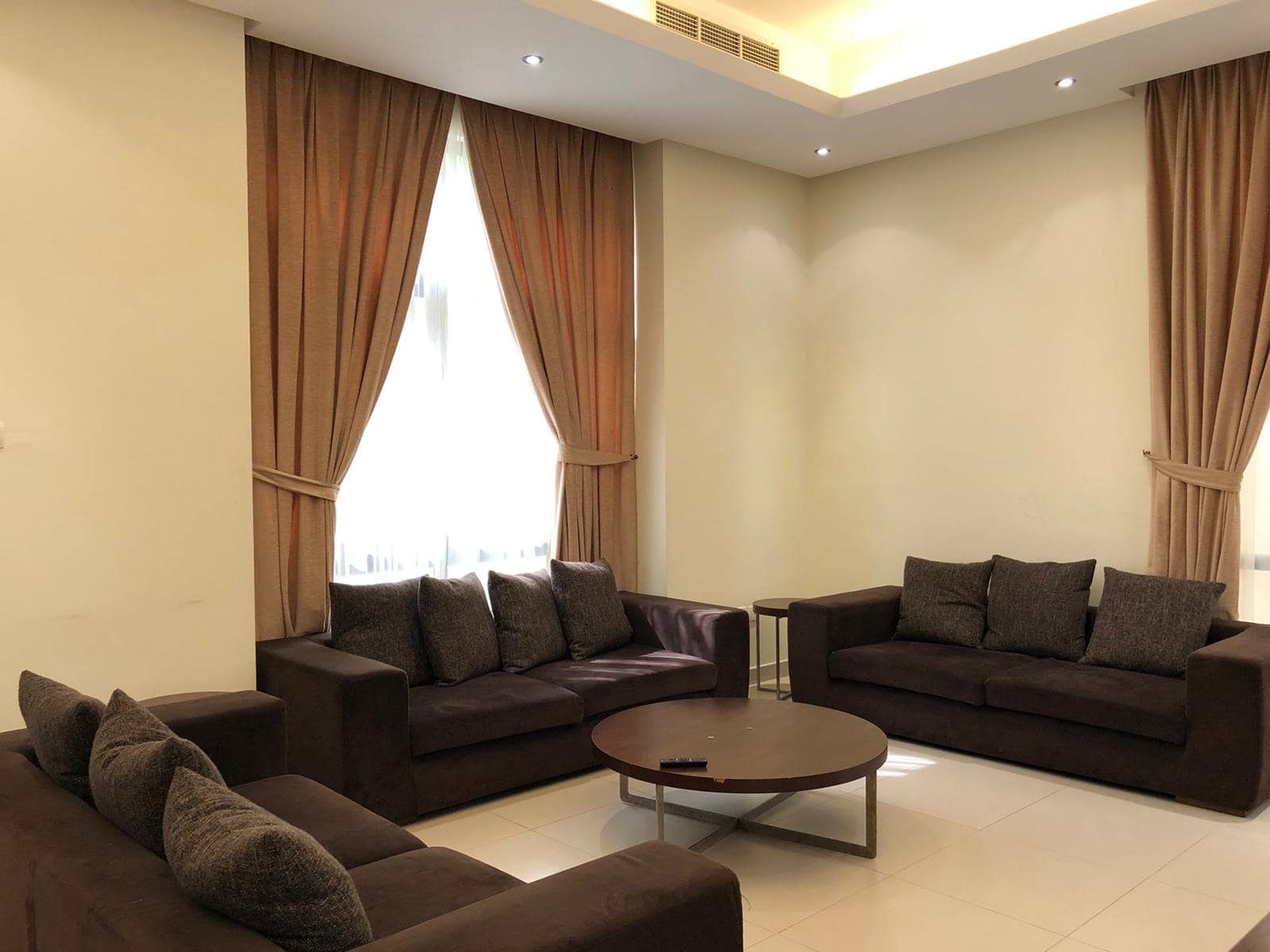 A living room with three brown sofas, a round wooden coffee table, and two windows with beige curtains. The room has tiled floors and ceiling lights.