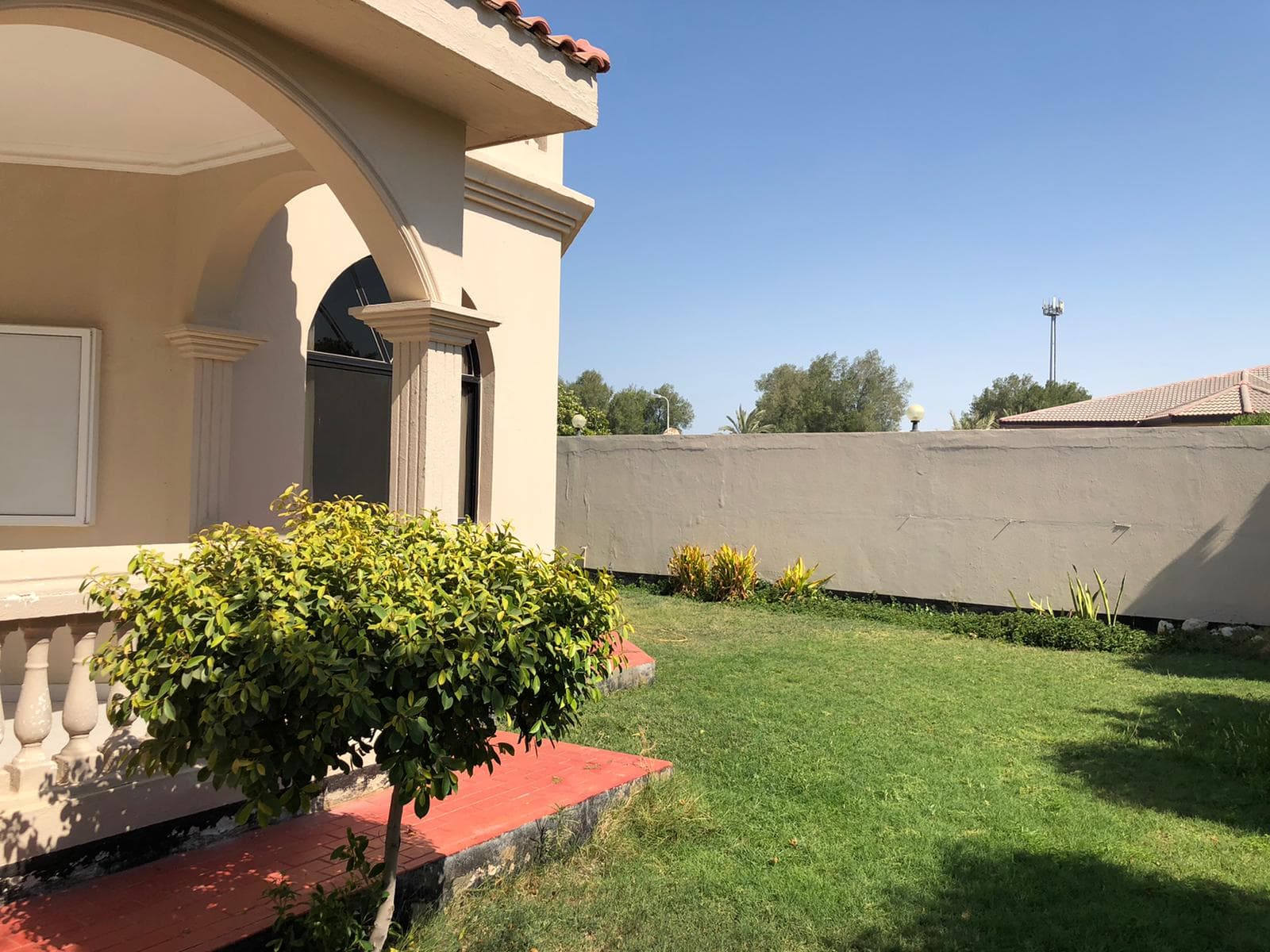 A sunny outdoor scene featuring a portion of a beige building with an archway, a green lawn, shrubs, and a tall boundary wall in the background.