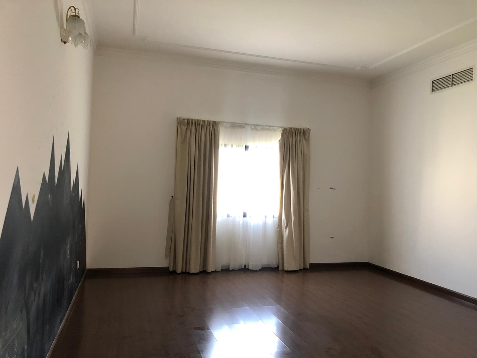 An empty room with pale walls, a small curtained window, and dark wooden flooring. One wall features a painted dark mountain design.