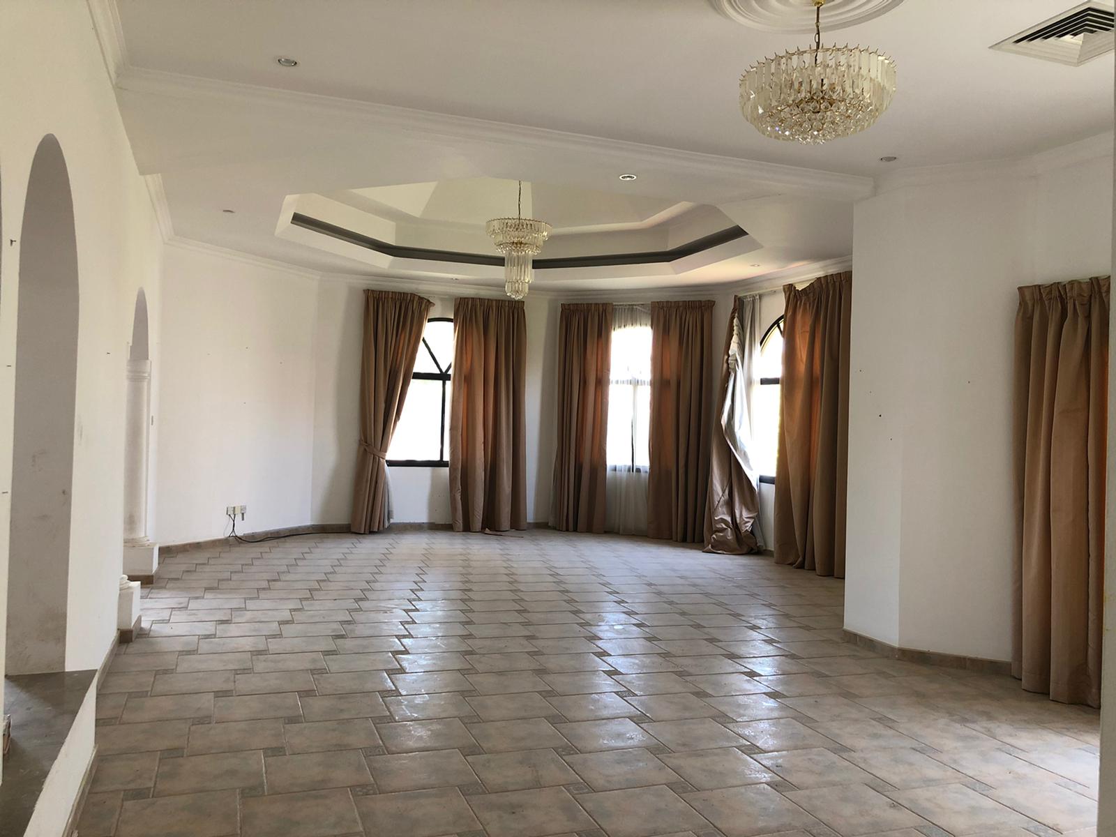 A spacious, empty room with tiled flooring, large windows with drawn beige curtains, and two ceiling chandeliers. The walls are white, and the room has a slightly elevated platform area on one side.