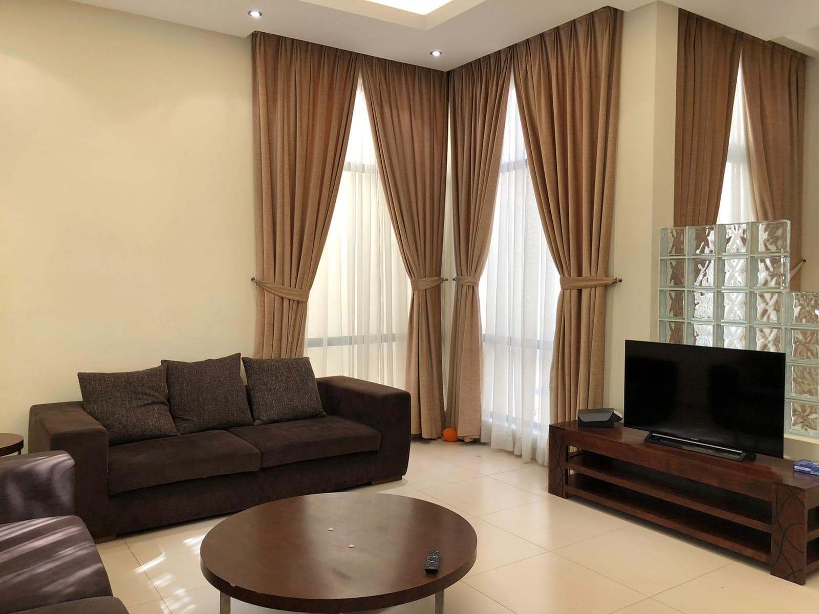 A living room with brown curtains, a dark brown sofa, a round wooden coffee table, and a wooden TV stand with a flat-screen TV. Bright neutral walls and large windows add light to the space.