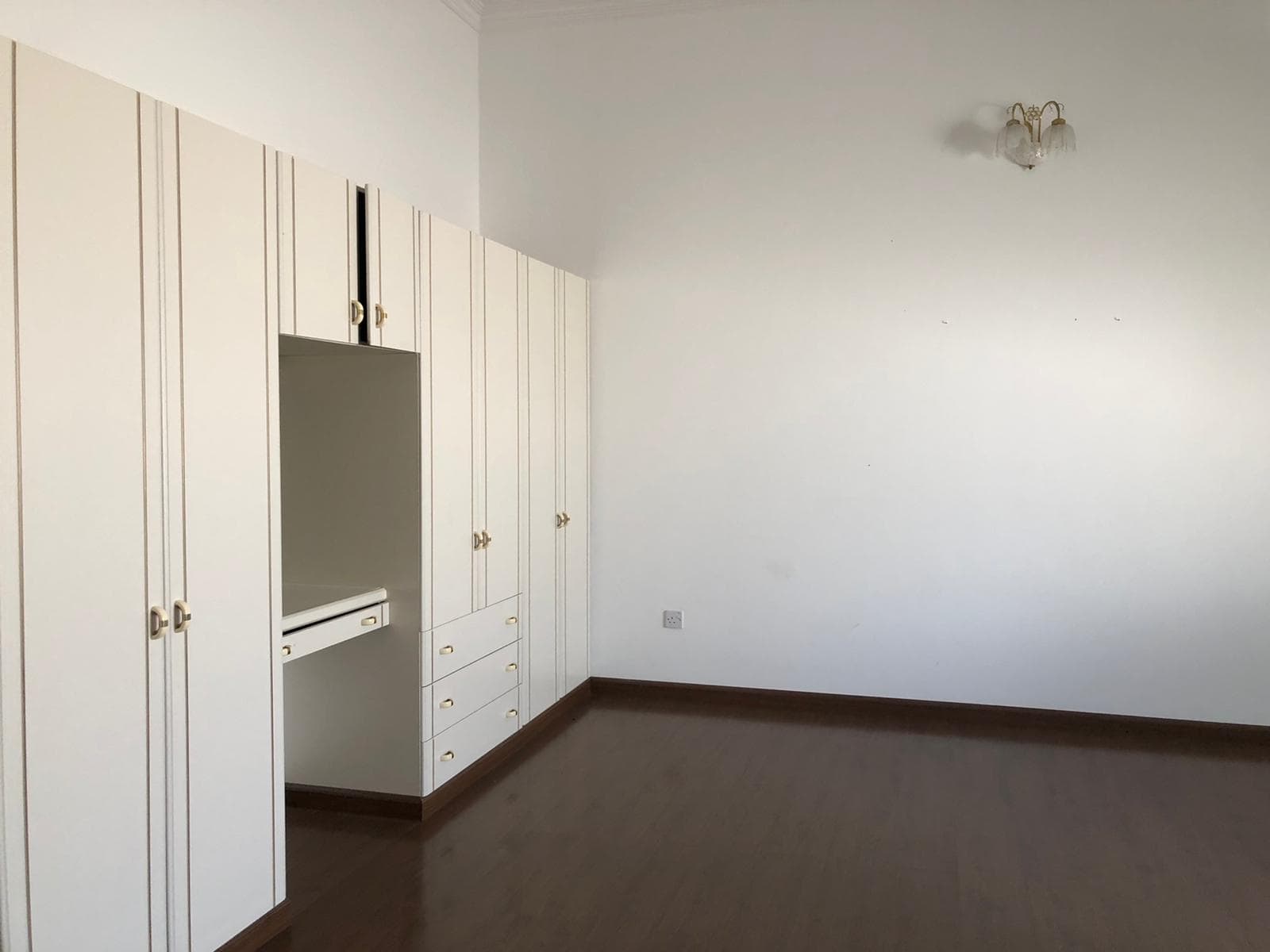 A room with white built-in wardrobes and drawers against a wall, wooden flooring, and an unlit wall sconce. The room is empty and well-lit.