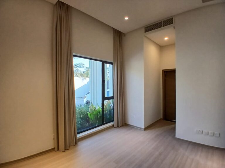 A minimalist, empty room with large window dressed with beige curtains. Room has wooden flooring, white walls, and a closed wooden door.