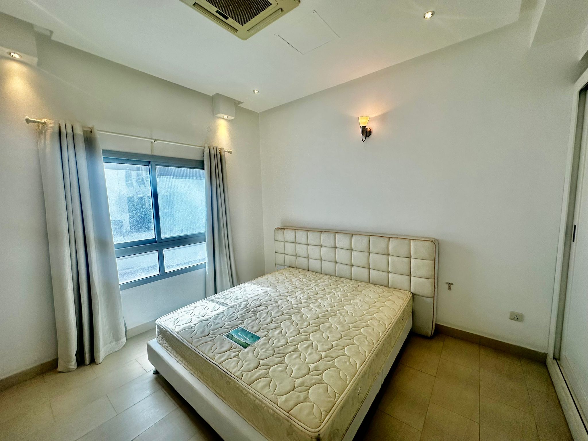 A minimally furnished bedroom with a large window, white walls, a double bed with a patterned mattress, and white curtains.