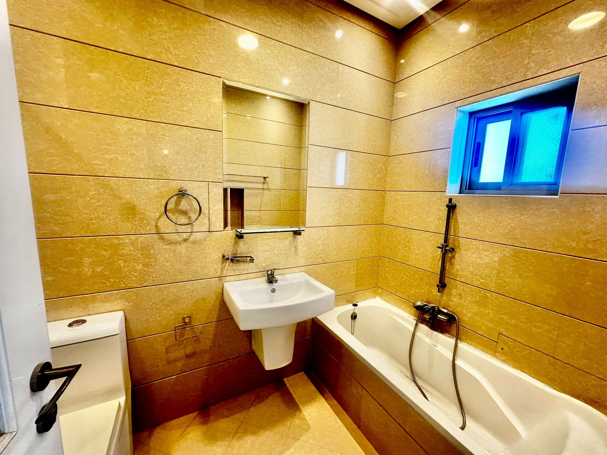 Modern bathroom with light beige tiles, a wall-mounted sink, toilet, bathtub with a shower attachment, rectangular mirror, and a small window.