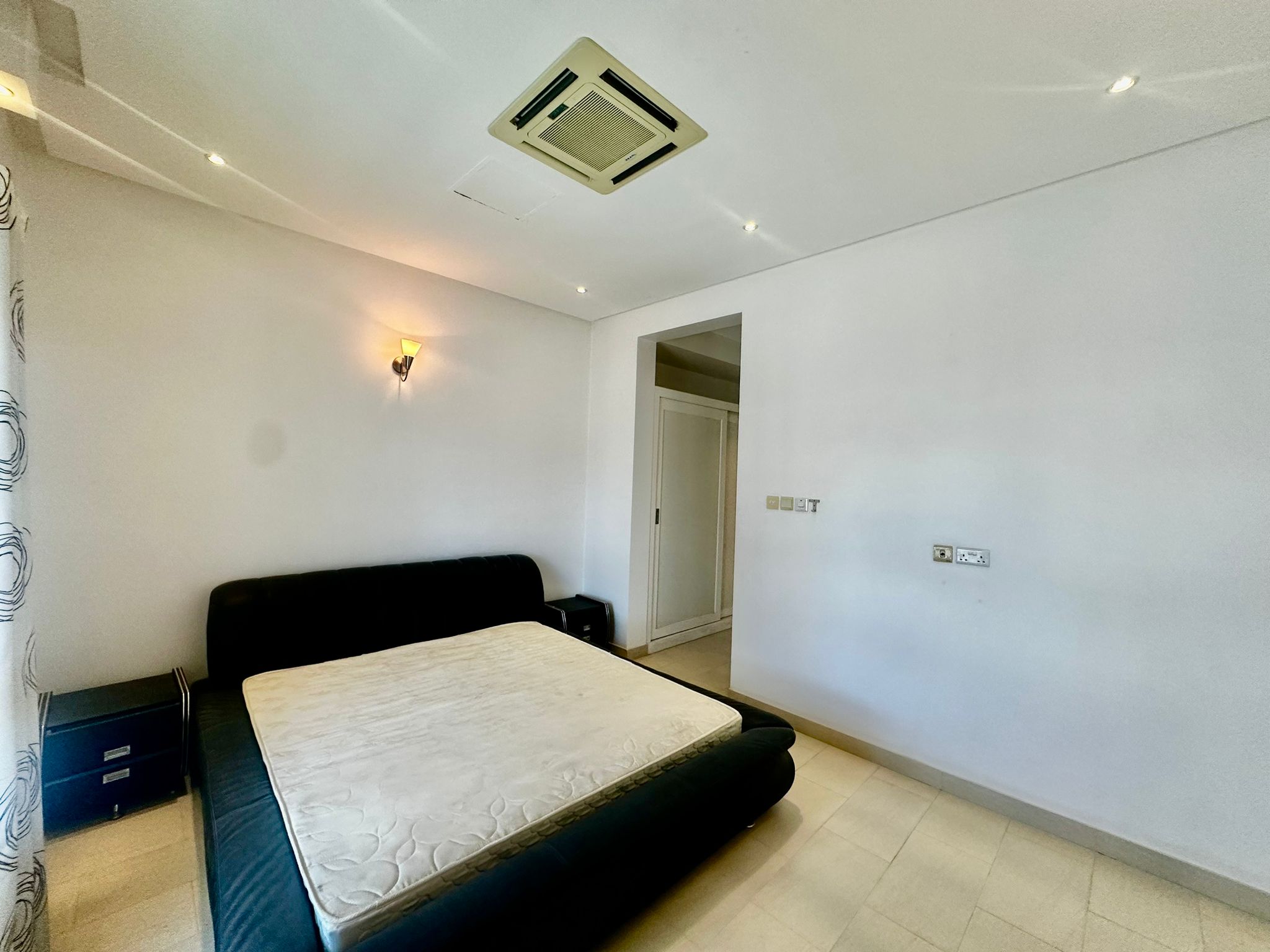 Minimalist bedroom with a black bed frame and mattress, bedside table with drawers, wall-mounted lamp, and an air conditioning unit on the ceiling. White walls and tiled floor. Closet visible in corner.