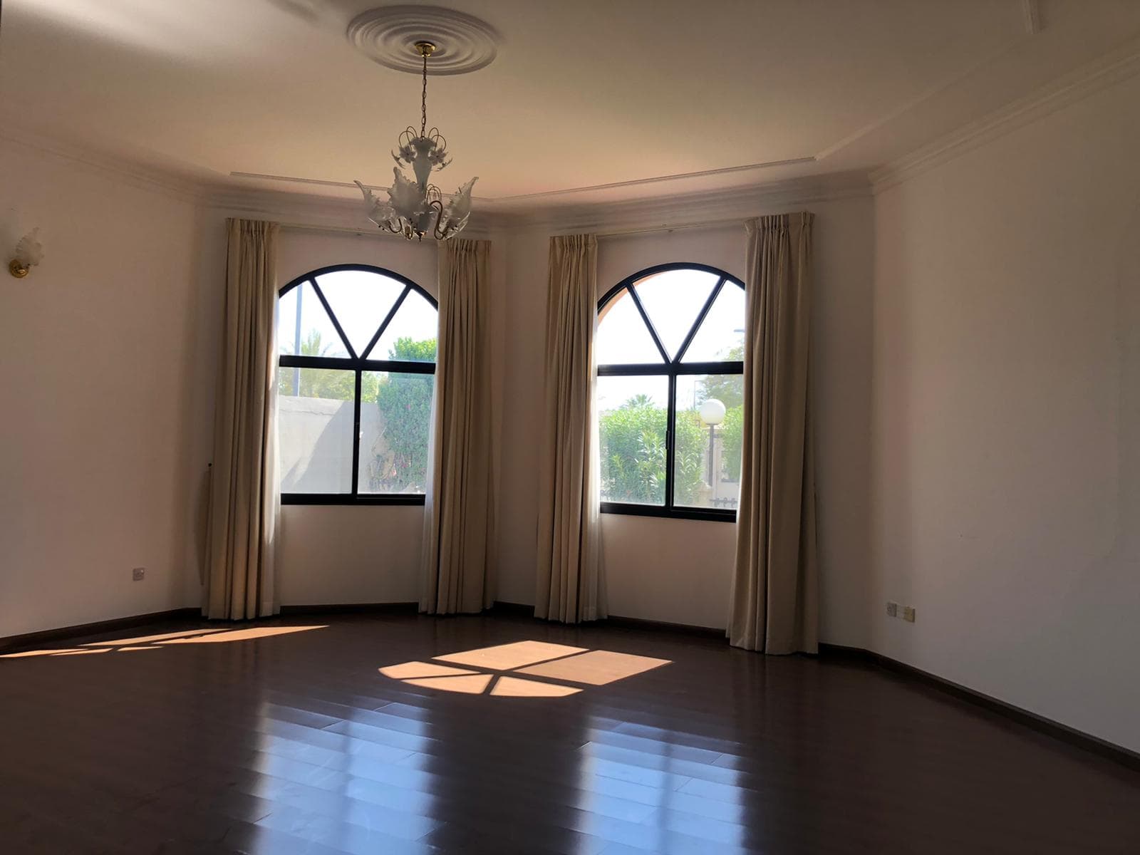 A spacious, sunlit room with dark wooden floors, arched windows with curtains, and a chandelier hanging from the ceiling.