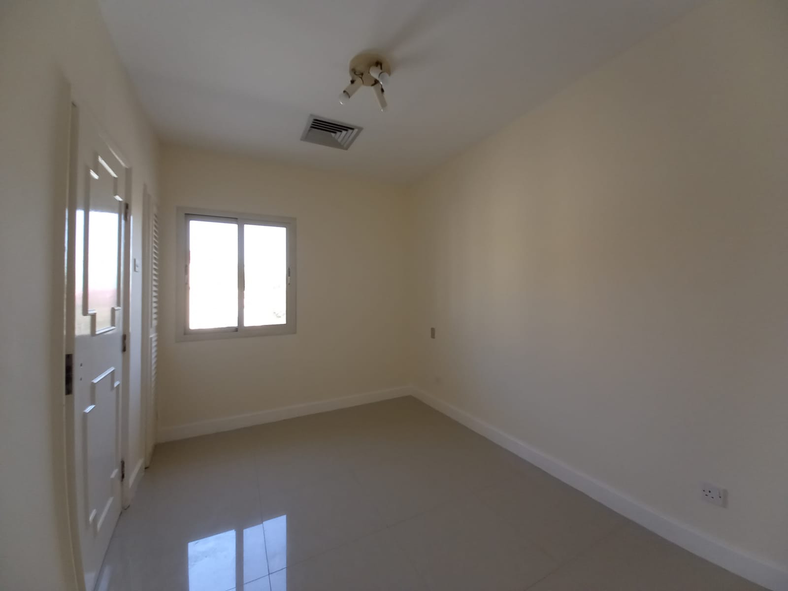 An empty, unfurnished room with a ceiling light, a window, a door, and tiled floor. The walls are painted beige, and the room is well-lit by natural light from the window.