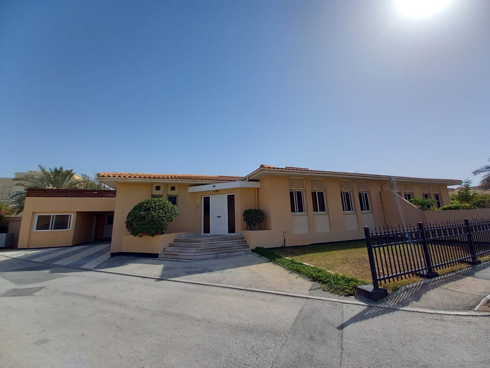 A single-story beige building with a flat roof and several windows stands under a clear blue sky. There is a staircase leading up to a white door, and a black fence borders the property.