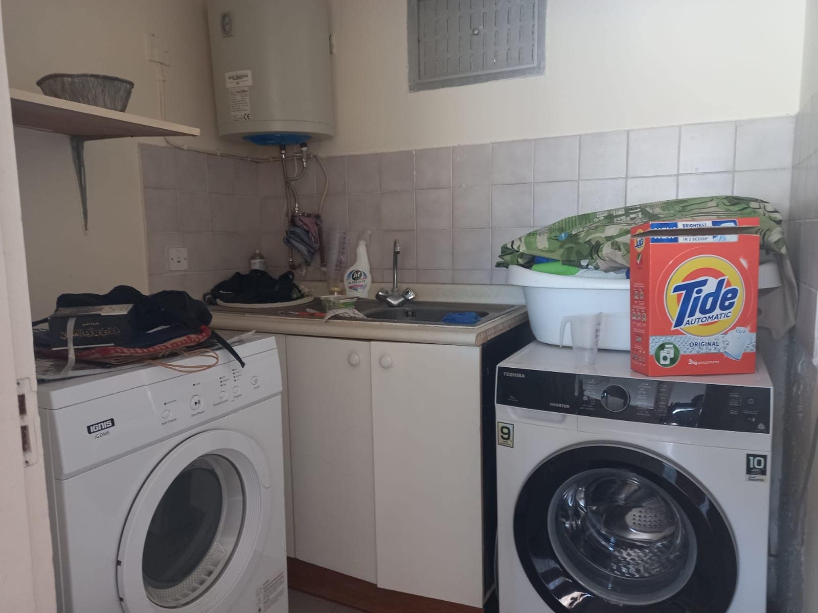 A laundry room with a washing machine, a dryer, a sink, shelves with various items, and a box of Tide detergent on the counter.