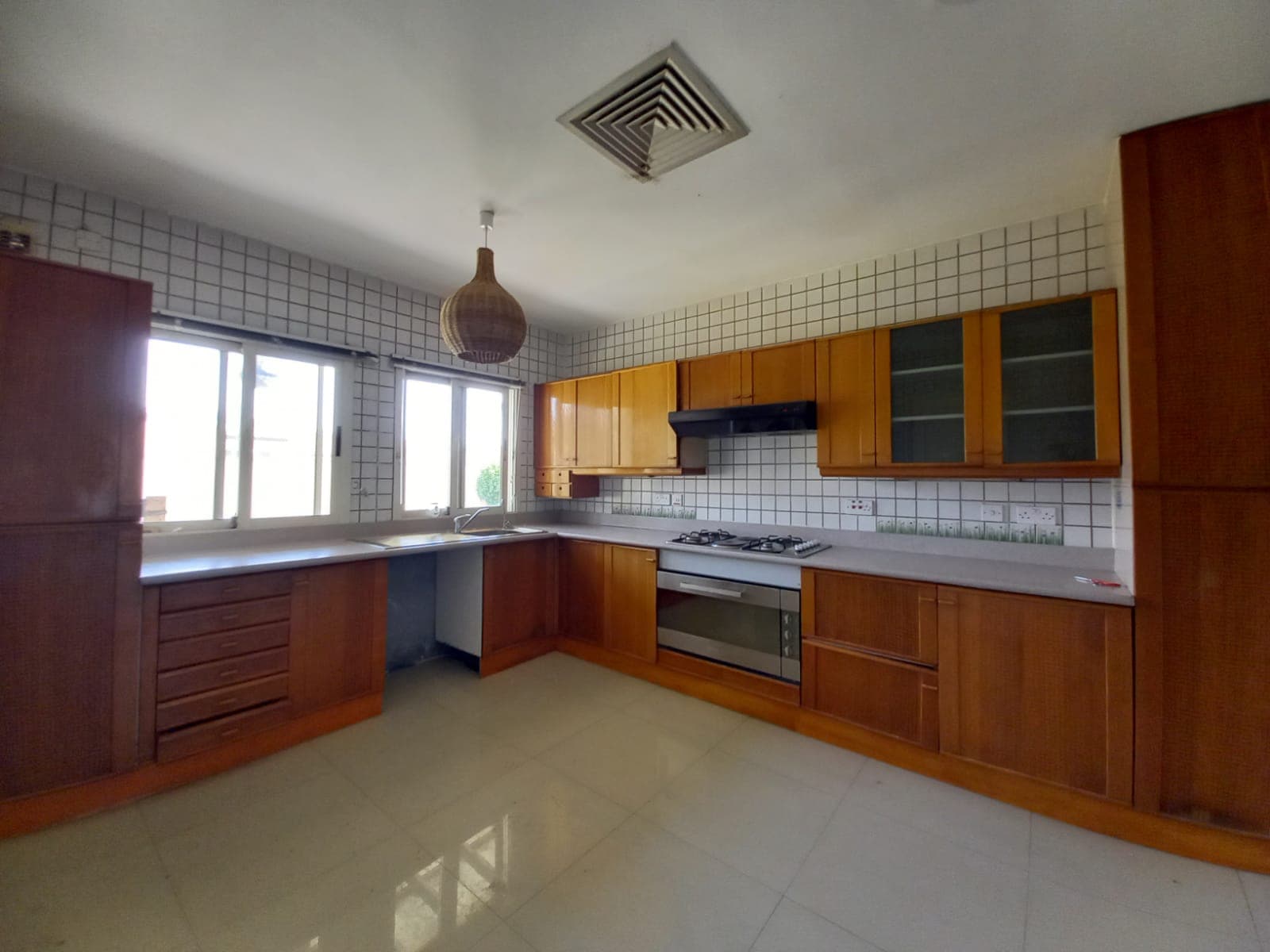 A spacious kitchen with wooden cabinets, white tiled walls, a built-in stovetop and oven, large windows, and a hanging wicker light fixture.