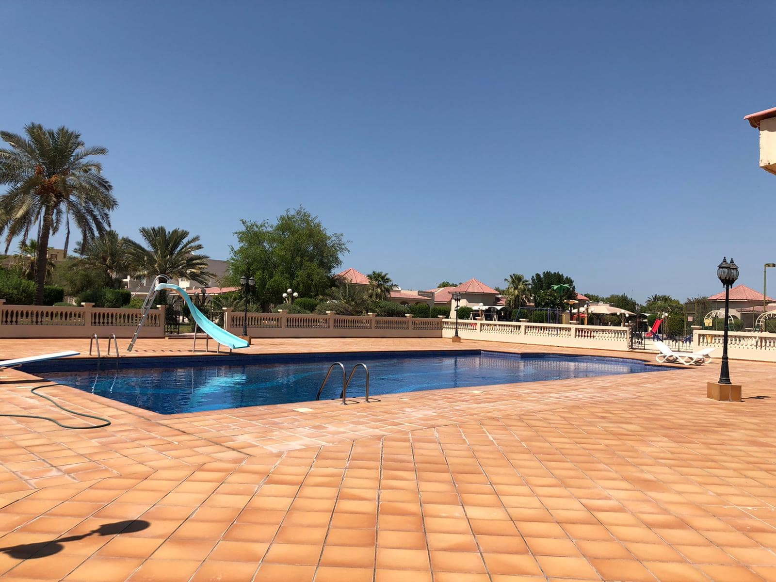 A rectangular outdoor swimming pool with a blue slide on one side, surrounded by a tiled patio. There are palm trees and houses with red roofs in the background.