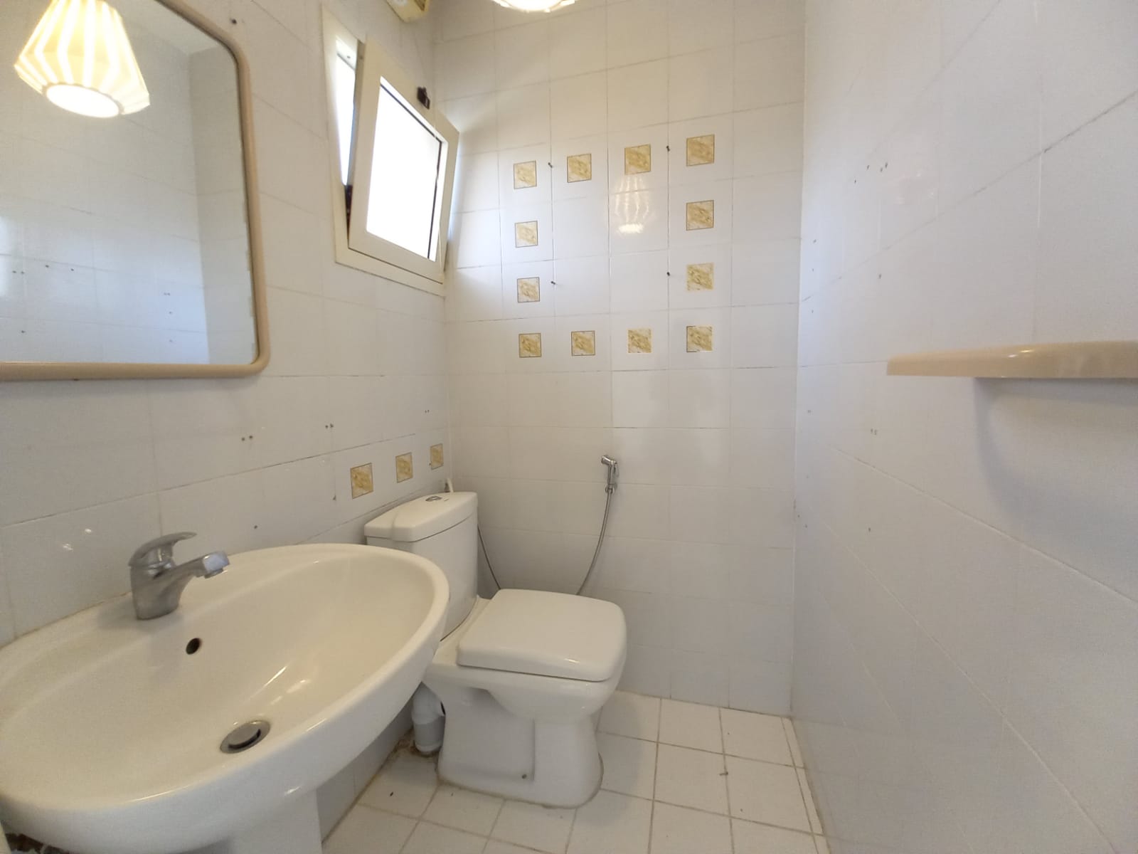 A small bathroom with white tiles, a sink with a faucet, a mirror, a toilet, and a bidet spray next to the toilet. A window near the ceiling provides natural light. A shelf is mounted on the wall.