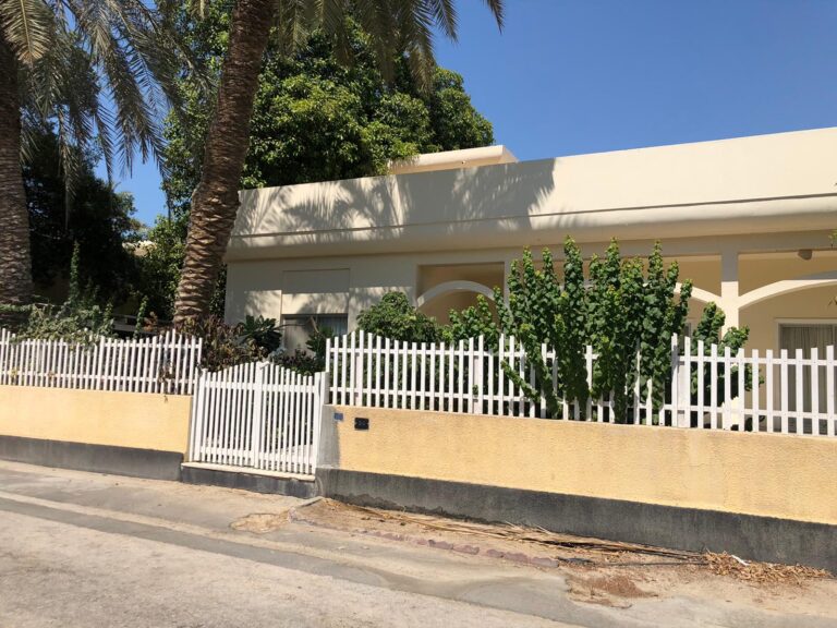 A beige house with a white picket fence, palm trees, and bushes in front. The property is situated along a clear road under a bright blue sky.